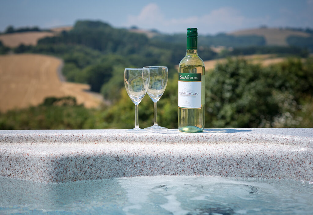 Two wine glasses and a bottle of white wine sat on the edge of a hot tub, looking out at hills, fields and trees