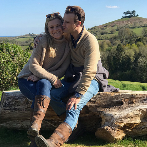 A man and a woman sat on a log hugging in front of a grassy hill